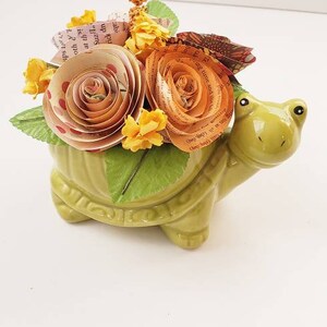 May include: A green ceramic turtle planter with a bouquet of paper flowers. The flowers are made from book pages and are in shades of orange, yellow, and pink.