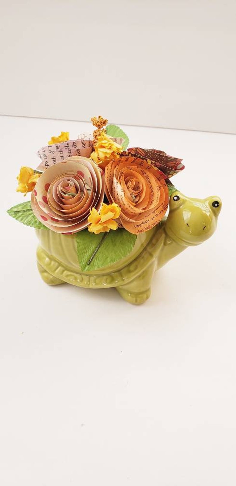 May include: A green ceramic turtle planter with a bouquet of paper flowers. The flowers are made from book pages and are in shades of orange, yellow, and pink.