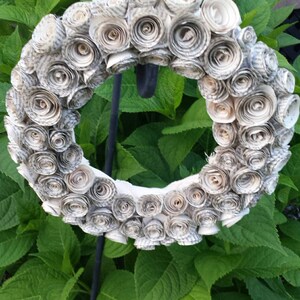 K&ouml;nnte beinhalten: Ein kreisf&ouml;rmiger Kranz aus gerollten Papierrosen in neutralen Farbt&ouml;nen. Der Kranz ist auf einem schwarzen Metallst&auml;nder montiert, vor dem Hintergrund &uuml;ppiger gr&uuml;ner Bl&auml;tter. Dieses dekorative Element ist f&uuml;r den Innen- und Au&szlig;enbereich geeignet.