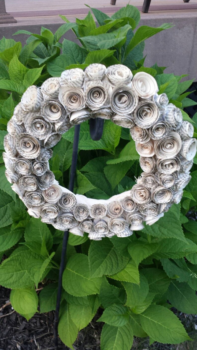 K&ouml;nnte beinhalten: Ein kreisf&ouml;rmiger Kranz aus gerollten Papierrosen in neutralen Farbt&ouml;nen. Der Kranz ist auf einem schwarzen Metallst&auml;nder montiert, vor dem Hintergrund &uuml;ppiger gr&uuml;ner Bl&auml;tter. Dieses dekorative Element ist f&uuml;r den Innen- und Au&szlig;enbereich geeignet.