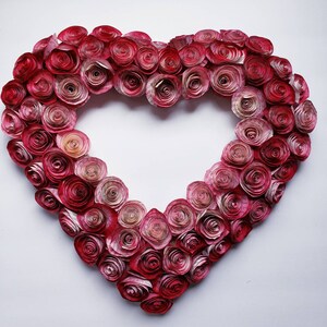 May include: A heart-shaped wreath made of paper roses in shades of pink and red. The roses are arranged in a gradient pattern, with the darkest roses at the center and the lightest roses at the edges.
