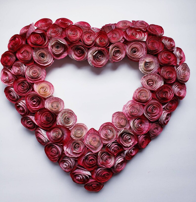 May include: A heart-shaped wreath made of paper roses in shades of pink and red. The roses are arranged in a gradient pattern, with the darkest roses at the center and the lightest roses at the edges.