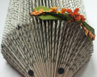 May include: A handmade hedgehog sculpture crafted from the pages of a book. The body is fan-shaped, with the pages forming the spines. It features black eyes and a nose, and is adorned with a green ribbon and orange floral accents.