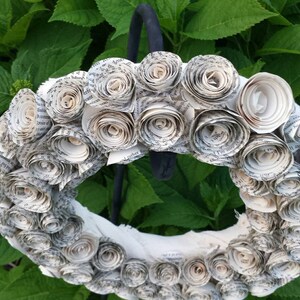 K&ouml;nnte beinhalten: Ein handgefertigter Kranz aus gerollten Papierrosen, der ein strukturiertes, florales Design erzeugt. Der Kranz ist wei&szlig; und cremefarben, mit sichtbarem Text aus dem Papier. Er wird vor einem Hintergrund aus &uuml;ppigen gr&uuml;nen Bl&auml;ttern pr&auml;sentiert.