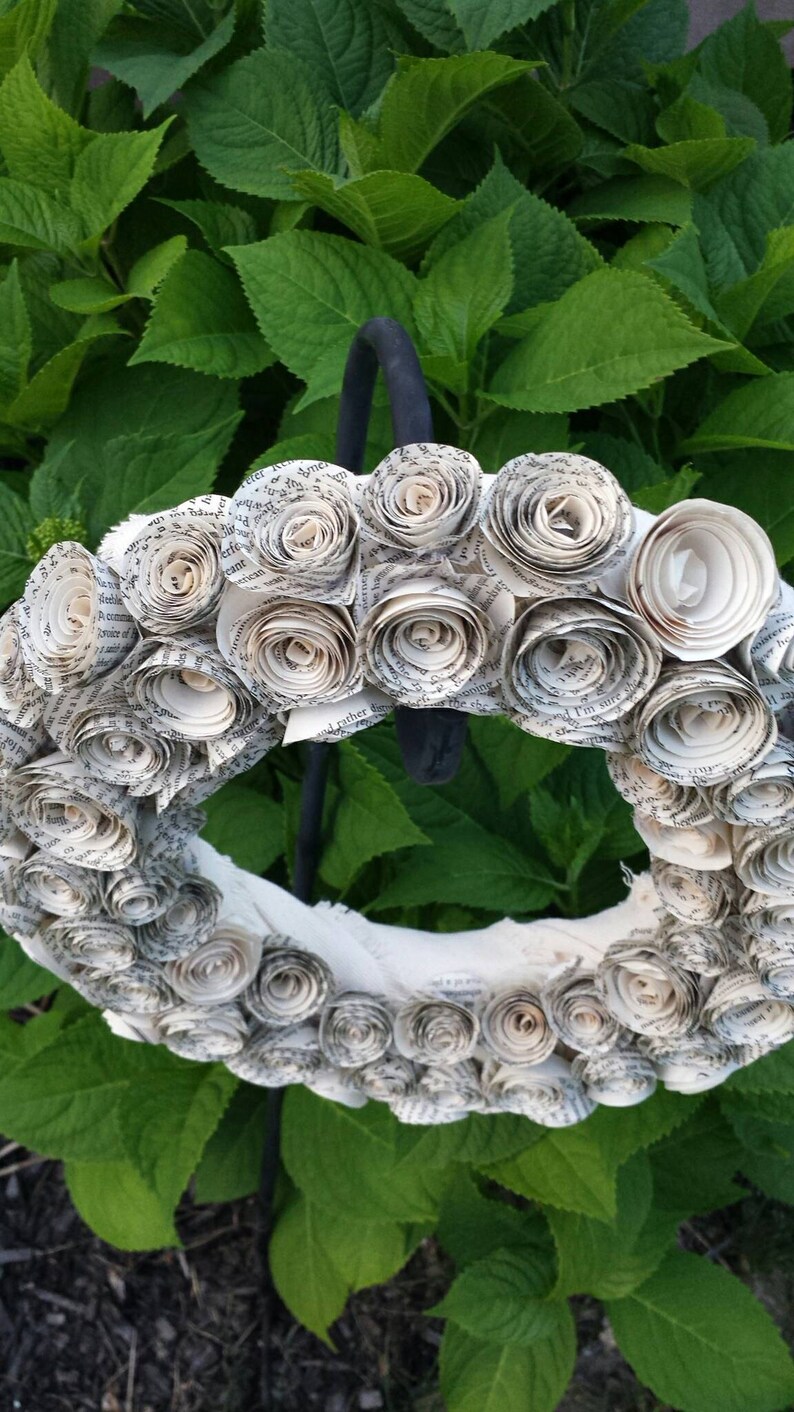 K&ouml;nnte beinhalten: Ein handgefertigter Kranz aus gerollten Papierrosen, der ein strukturiertes, florales Design erzeugt. Der Kranz ist wei&szlig; und cremefarben, mit sichtbarem Text aus dem Papier. Er wird vor einem Hintergrund aus &uuml;ppigen gr&uuml;nen Bl&auml;ttern pr&auml;sentiert.