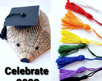 May include: A book folded into the shape of a graduation cap with a black tassel. The cap is decorated with a black graduation cap and a black tassel. The text "Celebrate 2022" is printed below the book. The image also shows a variety of colorful tassels.