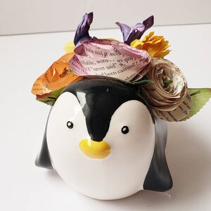 May include: A ceramic penguin planter with a black and white design. The planter is decorated with a colorful flower arrangement made of paper and fabric.