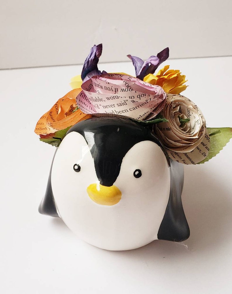 May include: A ceramic penguin planter with a black and white design. The planter is decorated with a colorful flower arrangement made of paper and fabric.