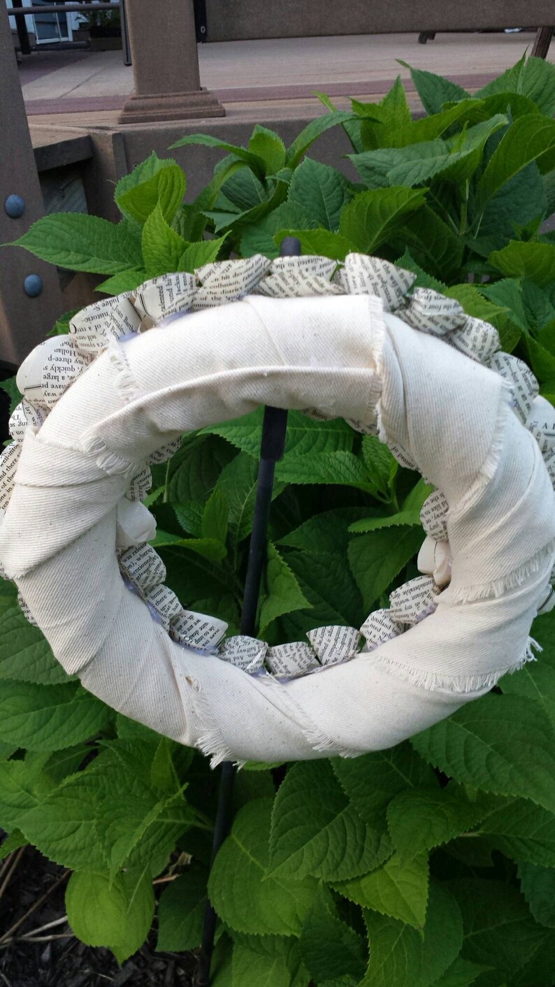 K&ouml;nnte beinhalten: Ein handgefertigter Kranz mit einer wei&szlig;en Stoffbasis und einem Rand aus gerolltem Papier. Der Kranz wird vor einem Hintergrund aus gr&uuml;nen Bl&auml;ttern pr&auml;sentiert. Der Kranz ist ein dekorativer Artikel, der sich f&uuml;r die Inneneinrichtung eignet.