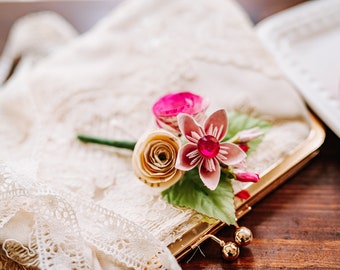May include: A small bouquet of paper flowers in shades of pink and cream, with a single pink rhinestone in the center of the largest flower. The bouquet is resting on a white lace fabric.