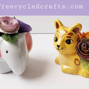 May include: Two ceramic planters, one shaped like an elephant and the other like a cat, are decorated with paper flowers. The elephant planter is white with pink ears and has purple and pink paper flowers. The cat planter is yellow with brown stripes and has orange and multi-colored paper flowers.