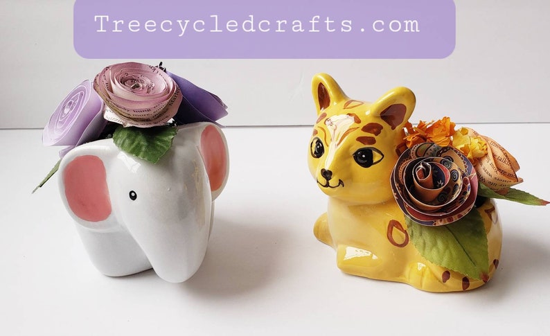 May include: Two ceramic planters, one shaped like an elephant and the other like a cat, are decorated with paper flowers. The elephant planter is white with pink ears and has purple and pink paper flowers. The cat planter is yellow with brown stripes and has orange and multi-colored paper flowers.