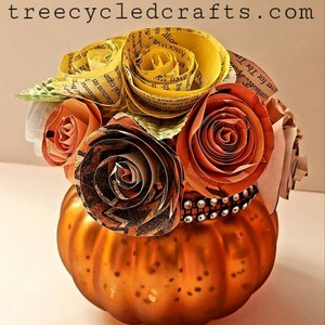May include: A small orange pumpkin vase with a decorative band of rhinestones around the top. The vase is filled with a bouquet of paper flowers made from book pages. The flowers are in shades of yellow, orange, and brown.