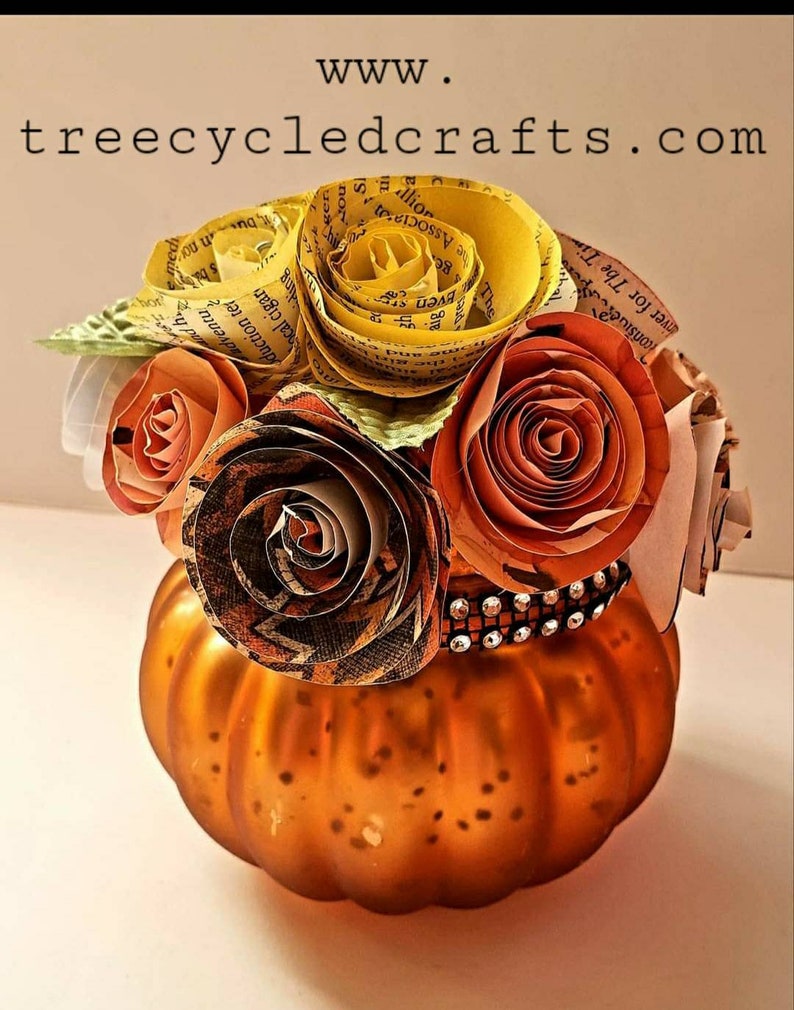 May include: A small orange pumpkin vase with a decorative band of rhinestones around the top. The vase is filled with a bouquet of paper flowers made from book pages. The flowers are in shades of yellow, orange, and brown.