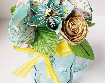 May include: A bouquet of paper flowers in a blue mason jar. The flowers are made from book pages and are decorated with bees. The bouquet is tied with a yellow ribbon.