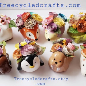May include: Eight ceramic animal planters with paper flowers. The planters are shaped like a fox, hedgehog, turtle, elephant, penguin, walrus, and two white bears. The flowers are made of colorful paper and are arranged in a bouquet style. The planters are all different colors and have a variety of designs. Treecycledcrafts.com and Treecycledcrafts.etsy.com are printed on the white surface.
