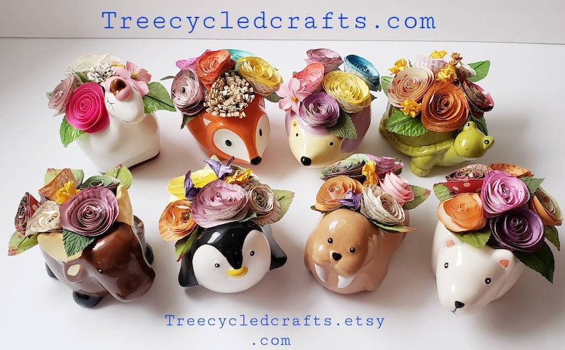 May include: Eight ceramic animal planters with paper flowers. The planters are shaped like a fox, hedgehog, turtle, elephant, penguin, walrus, and two white bears. The flowers are made of colorful paper and are arranged in a bouquet style. The planters are all different colors and have a variety of designs. Treecycledcrafts.com and Treecycledcrafts.etsy.com are printed on the white surface.