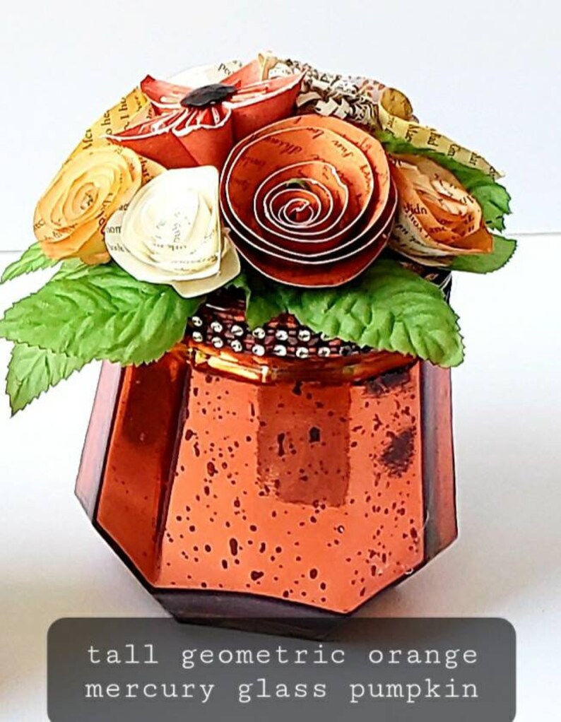May include: Tall geometric orange mercury glass pumpkin vase with a bouquet of paper flowers. The flowers are made from book pages and are in shades of orange, yellow, and white.