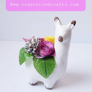 May include: White ceramic llama planter with a brown glaze, decorated with paper flowers made from book pages in shades of pink and yellow. The flowers are accented with green leaves.