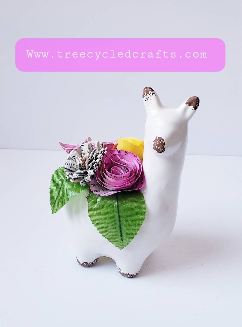 May include: White ceramic llama planter with a brown glaze, decorated with paper flowers made from book pages in shades of pink and yellow. The flowers are accented with green leaves.