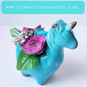 May include: A turquoise ceramic unicorn figurine with a pink paper flower made from book pages. The unicorn has a brown horn and brown accents on its hooves. The flower has a brown wire stem.