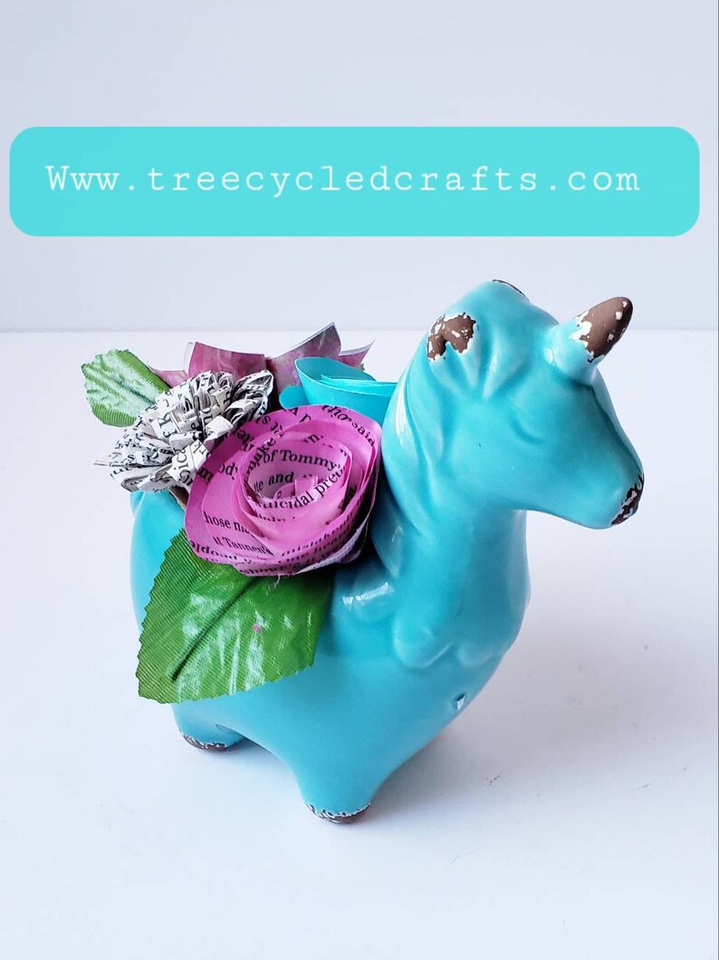 May include: A turquoise ceramic unicorn figurine with a pink paper flower made from book pages. The unicorn has a brown horn and brown accents on its hooves. The flower has a brown wire stem.