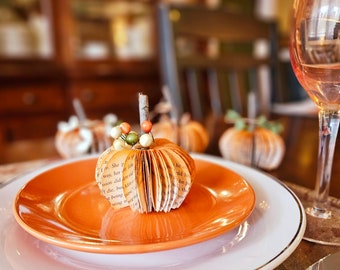 May include: A table setting with a white plate, an orange plate, and a book page pumpkin centerpiece. The centerpiece is decorated with small red and green beads. A wine glass and a silver knife are also on the table.