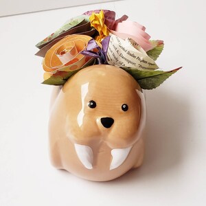 May include: A ceramic walrus planter with a white glaze and black eyes. The planter is decorated with a bouquet of paper flowers in shades of pink, orange, yellow, and purple.