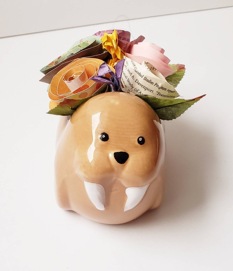 May include: A ceramic walrus planter with a white glaze and black eyes. The planter is decorated with a bouquet of paper flowers in shades of pink, orange, yellow, and purple.