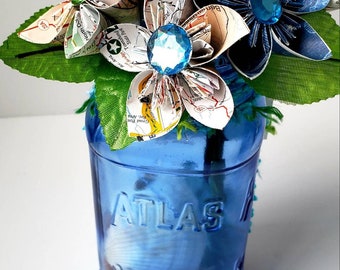 May include: A bouquet of paper flowers made from maps in a blue glass mason jar. The flowers are blue, white, and green. The jar has the word "ATLAS" embossed on it.