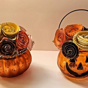 May include: Two orange glass pumpkin-shaped vases with paper flowers. The vase on the right has a black handle. The flowers are made of paper and are in shades of yellow, orange, and brown.