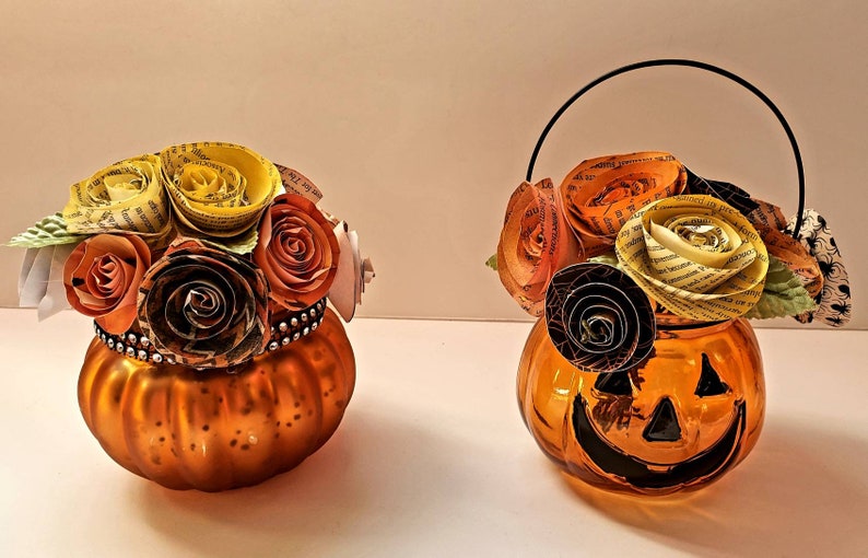 May include: Two orange glass pumpkin-shaped vases with paper flowers. The vase on the right has a black handle. The flowers are made of paper and are in shades of yellow, orange, and brown.