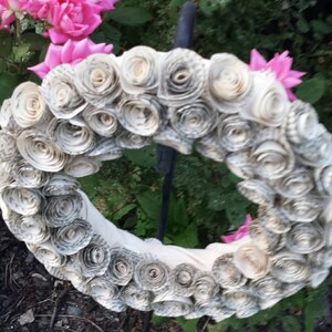 K&ouml;nnte beinhalten: Ein handgefertigter Kranz aus gerollten Buchseiten, der ein kreisf&ouml;rmiges Design bildet. Der Kranz ist mit Papierrosen geschm&uuml;ckt und vor dem Hintergrund von rosa Rosen und Gr&uuml;n angeordnet, was ein rustikales, florales Thema schafft.