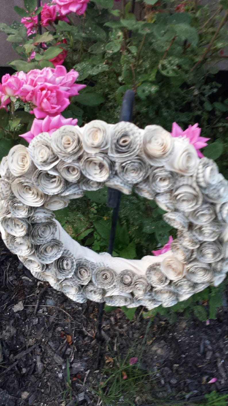 K&ouml;nnte beinhalten: Ein handgefertigter Kranz aus gerollten Buchseiten, der ein kreisf&ouml;rmiges Design bildet. Der Kranz ist mit Papierrosen geschm&uuml;ckt und vor dem Hintergrund von rosa Rosen und Gr&uuml;n angeordnet, was ein rustikales, florales Thema schafft.