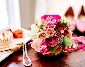 May include: A pink and peach paper flower bouquet with green leaves. The bouquet is on a wooden table with a white plate, a gold crown, and a pearl necklace.