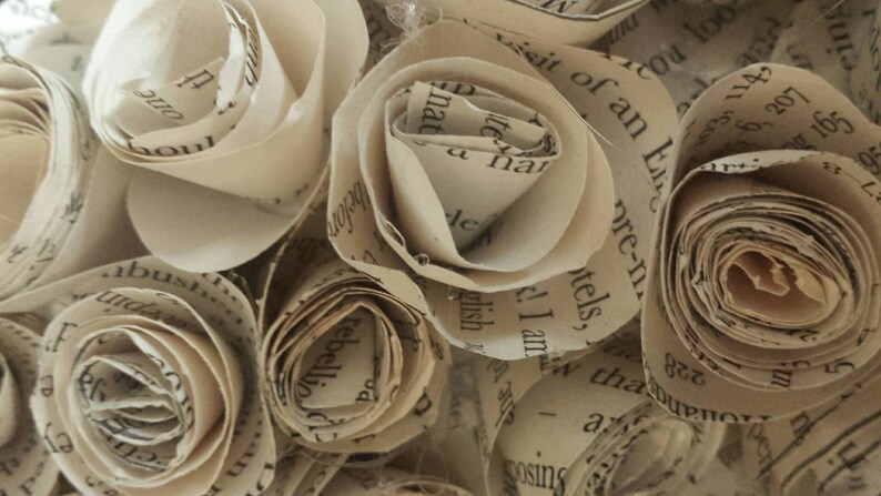 K&ouml;nnte beinhalten: Nahaufnahme von Papierrosen, jede sorgf&auml;ltig aus Buchseiten gefertigt. Die Rosen befinden sich in verschiedenen Bl&uuml;hstadien und zeigen die zarte Textur und den gedruckten Text. Die Farbe ist ein warmes, gealtertes Beige.
