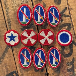 May include: A collection of ten embroidered fabric patches arranged in three rows. Five oval patches feature a white monument and a red sword on a blue background. Two octagonal patches are red with white crosses, one is a white flower with a blue star, and one is a circular red, white, and blue emblem.