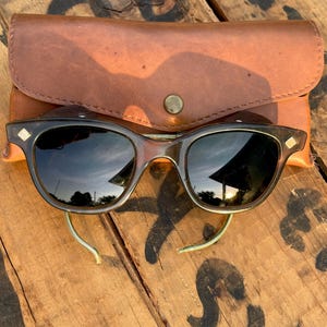 May include: Dark brown translucent sunglasses with a rounded frame, silver-toned diamond-shaped accents at the hinges, and curved metal temple tips, accompanied by a tan leather protective case featuring a single brass-colored snap button closure.