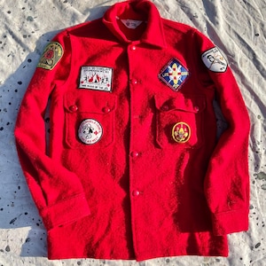 May include: A bright red long-sleeve wool shirt featuring a button-down front, two chest pockets, and multiple embroidered patches. The patches include a Snoopy graphic, a Life Guard emblem, and text reading Area 20 Conclave Crumhorn '83.