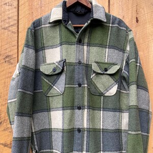 May include: A long-sleeve button-up shirt featuring a plaid pattern in shades of olive green, dark grey, and off-white. The shirt includes a pointed collar, two chest pockets with buttoned flaps, and black buttons down the front placket.