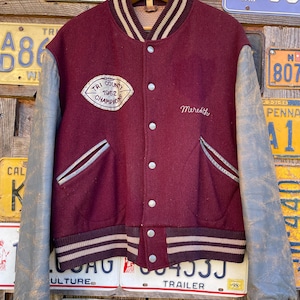 May include: A vintage maroon and gray varsity jacket with the text "Tri County 1962 Champions" and "Meredith" embroidered on the front. The jacket has gray leather sleeves, a striped collar, cuffs, and waistband, and a button-up closure.