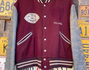May include: A vintage maroon and gray varsity jacket with the text "Tri County 1962 Champions" and "Meredith" embroidered on the front. The jacket has gray leather sleeves, a striped collar, cuffs, and waistband, and a button-up closure.