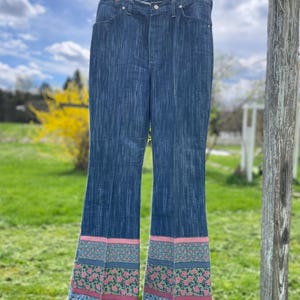 Pu&ograve; includere: Jeans blu a zampa con motivo floreale sull'orlo. Il motivo floreale comprende rose rosa, foglie verdi e uno sfondo azzurro. I jeans sono appesi a un filo.