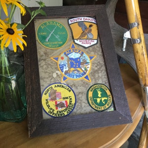 May include: A framed collection of embroidered patches featuring state and wildlife conservation organizations. The patches are in a dark wood frame and include organizations from Alabama, South Dakota, Alaska, Washington, and North America. The patches feature various wildlife, including a pheasant, a bear, and a deer.