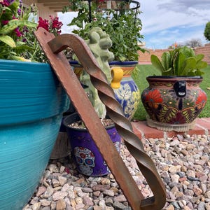 May include: A rusty brown, hand-forged metal door pull with a twisted center design. The handle is rectangular with a curved end. The background includes colorful flower pots and a gravel ground.