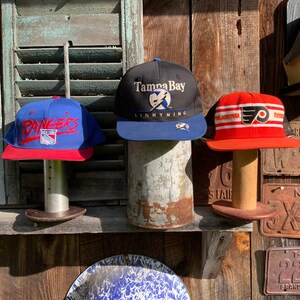 May include: Three vintage baseball caps on display. The left cap is blue with red trim and says "Rangers" in white. The middle cap is black with blue trim and says "Tampa Bay Lightning" in white. The right cap is red with white and black trim and says "Philadelphia Flyers" in white.