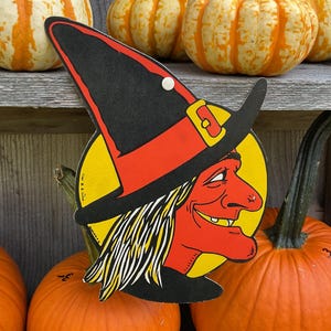 May include: A flat, die-cut cardboard decoration featuring a profile illustration of a witch with orange skin, a black pointed hat with a red band and yellow buckle, and yellow hair, set against a circular yellow background with a small white pin.