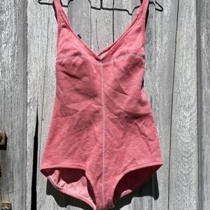 May include: A one-piece swimsuit in a muted red hue, hanging on a black hanger against a weathered wooden background. The swimsuit features a V-neckline with a crisscross design and white stitching details. The garment is made of a soft, textured fabric.