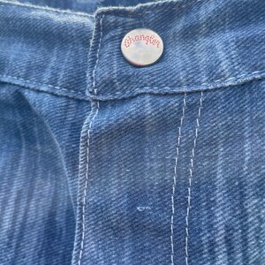 Pu&ograve; includere: Primo piano di jeans blu in denim con cuciture bianche. Il bottone argentato presenta il marchio "Wrangler" in lettere rosse. I jeans hanno un aspetto classico e usurato.