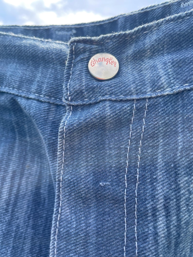 Pu&ograve; includere: Primo piano di jeans blu in denim con cuciture bianche. Il bottone argentato presenta il marchio "Wrangler" in lettere rosse. I jeans hanno un aspetto classico e usurato.